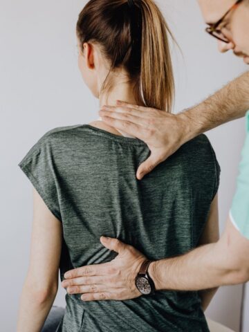 A healthcare professional examines a woman's upper back and neck, checking for spine symptoms while she sits facing away from the camera.