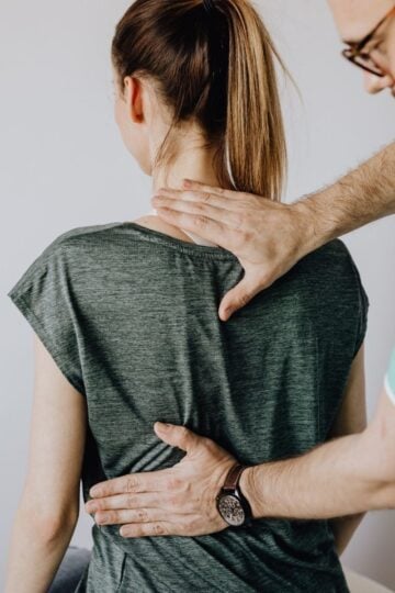A healthcare professional examines a woman's upper back and neck, checking for spine symptoms while she sits facing away from the camera.