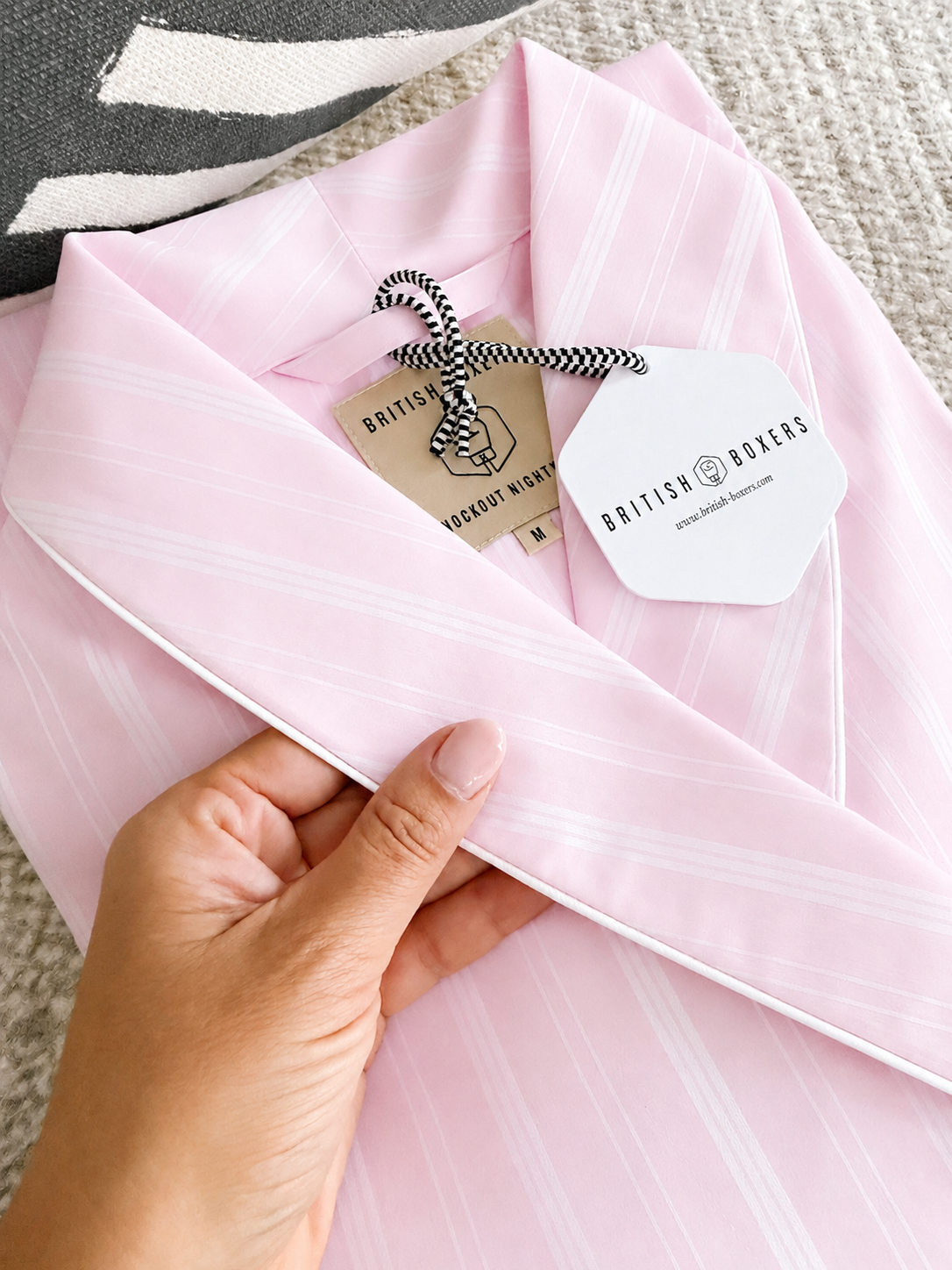 A hand holds the collar of a pink and white striped pyjama top with white piping, showing tags labelled "British Boxers" and "100% Cotton Nightwear.