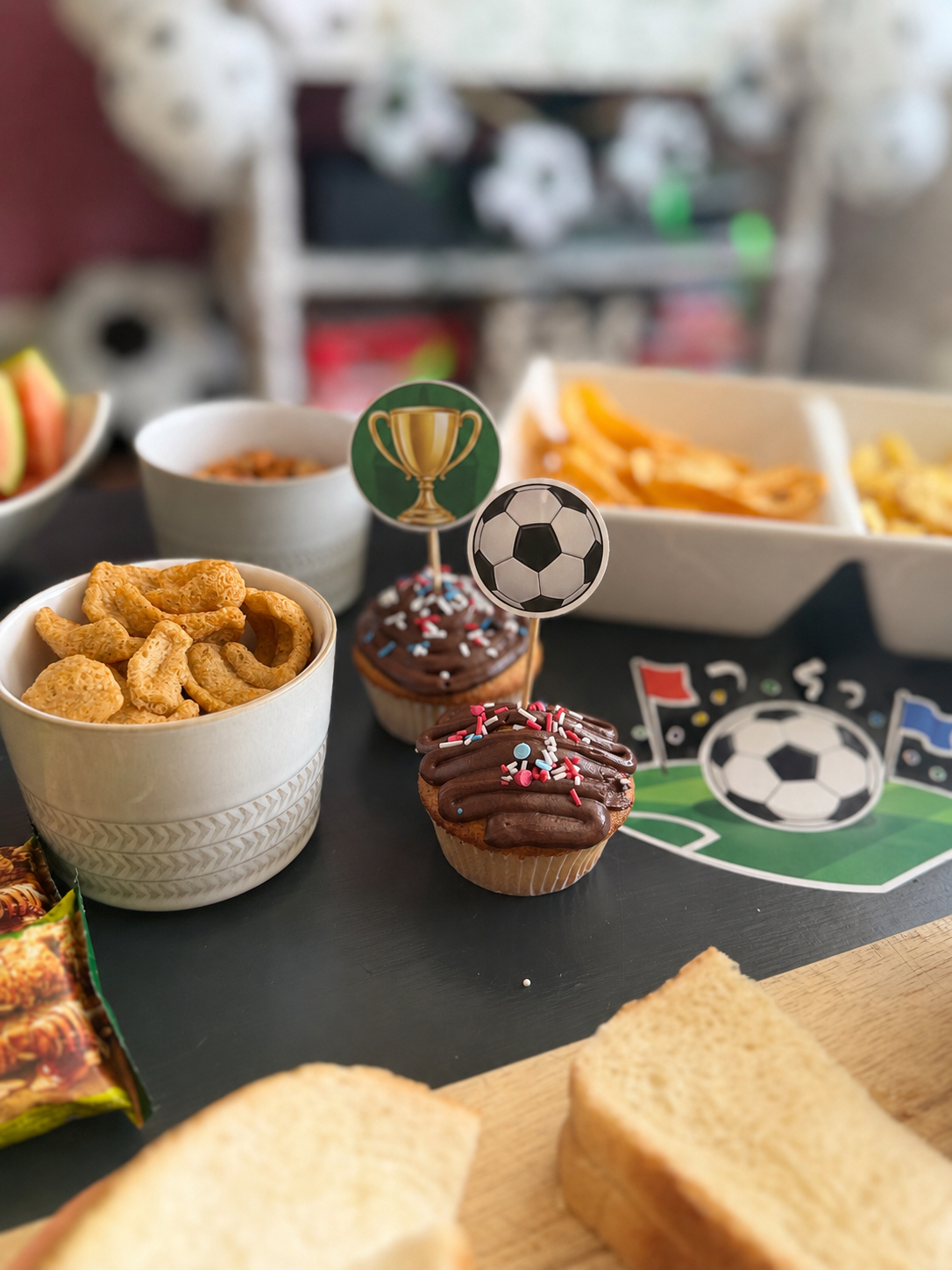 How To Host A Football Watch Party At Home This Summer A table set for a football watch party with snacks, cupcakes decorated with football-themed toppers, sliced bread, crisps, and a football-themed placemat in the background—perfect to host at home this summer.