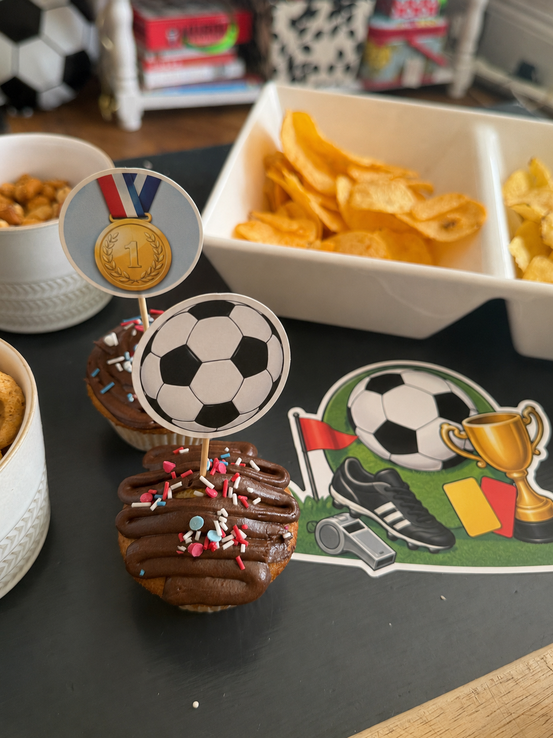 How To Host A Football Watch Party At Home This Summer Chocolate cupcakes with football-themed toppers and sprinkles are displayed on a tray next to a bowl of crisps—perfect treats to serve at home for your summer football watch party.