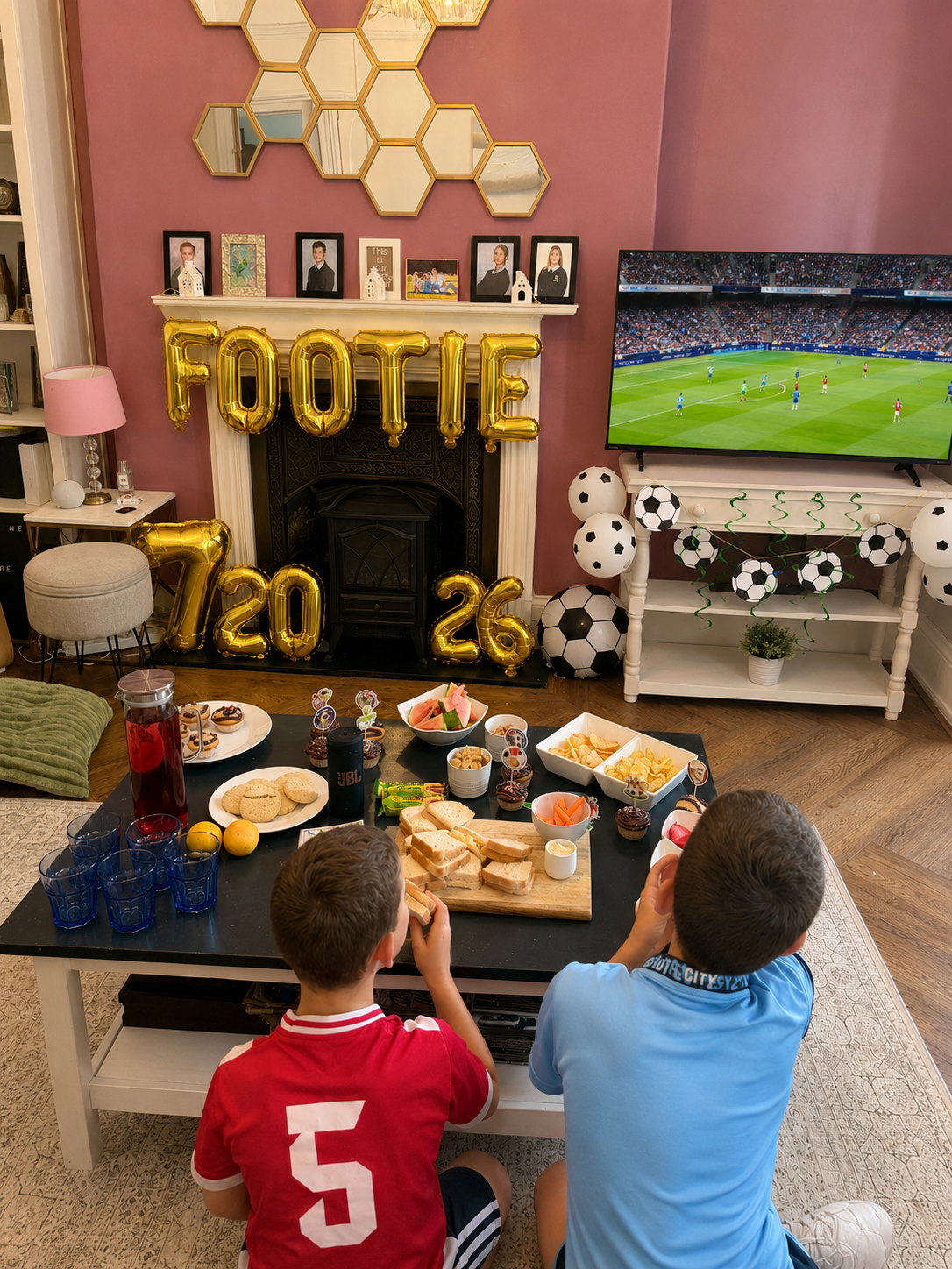 How To Host A Football Watch Party At Home This Summer Two children enjoy a summer football watch party in a living room decorated with gold “FOOTIE” balloons and football-themed decor, as they relax with snacks and drinks arranged on the table in front of them.