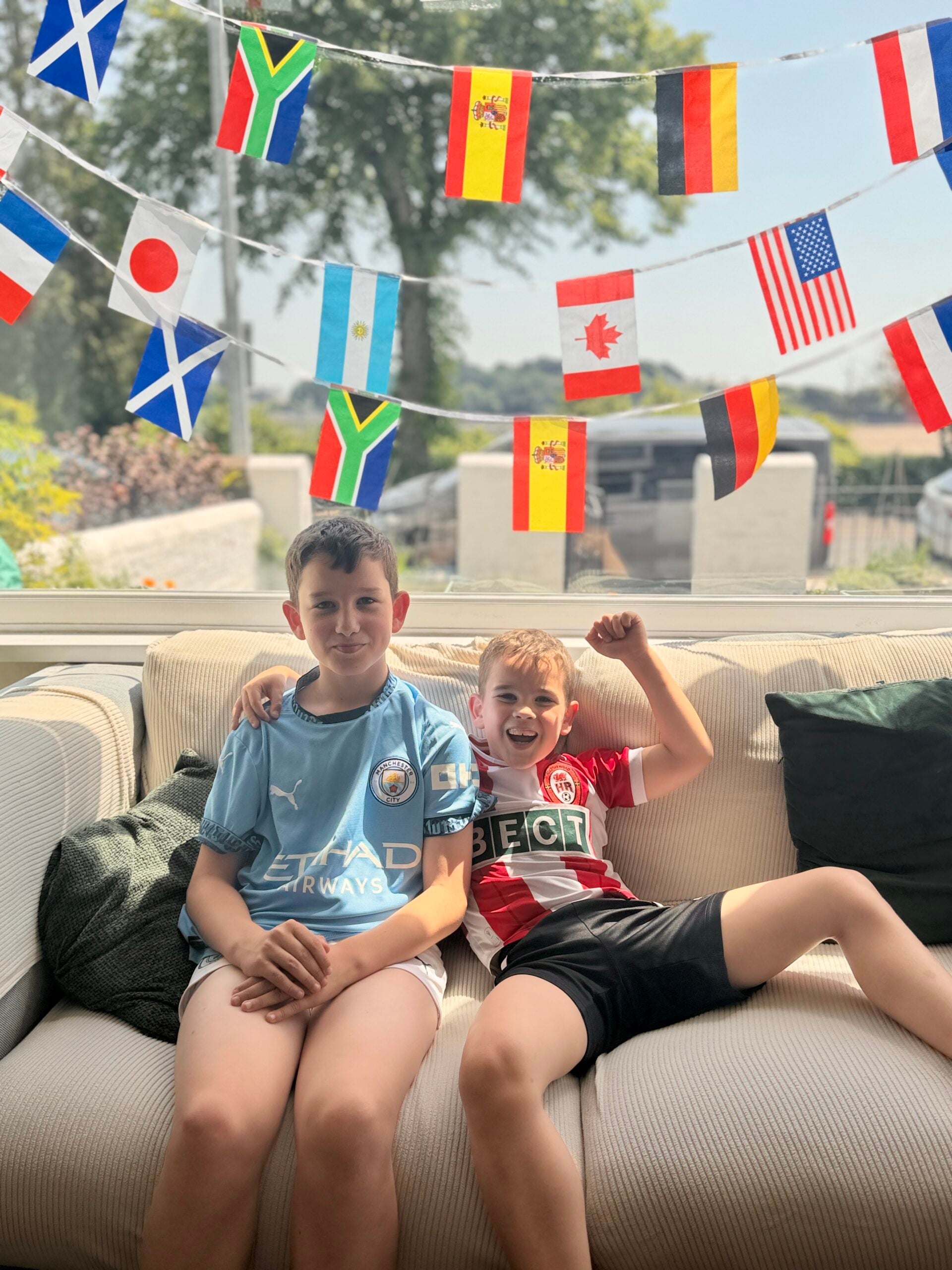 How To Host A Football Watch Party At Home This Summer Two boys sit on a sofa under a string of international flags. One wears a Manchester City football shirt, the other wears a red and white football shirt and is cheering with his arm raised.