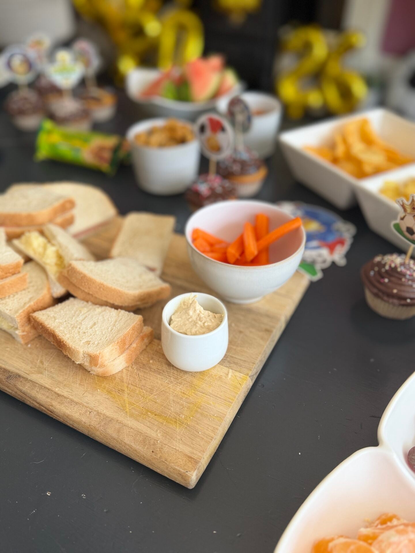 How To Host A Football Watch Party At Home This Summer A wooden board with stacked sandwiches and a small cup of dip, surrounded by bowls of carrot sticks, crisps, cupcakes, and other snacks on a dark table.