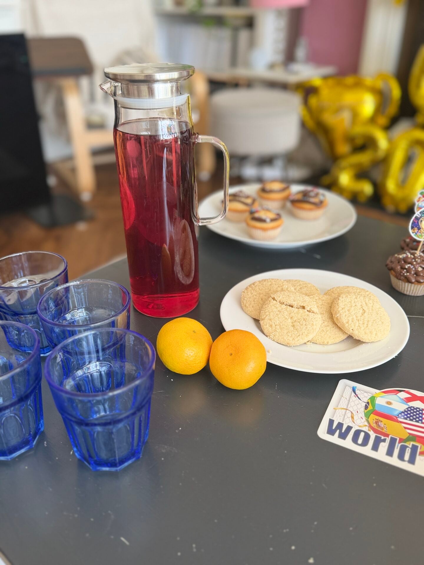 How To Host A Football Watch Party At Home This Summer A table with blue glasses, a jug of red drink, two oranges, a plate of biscuits, fairy cakes, and a "world" card, with balloons and snacks in the background.