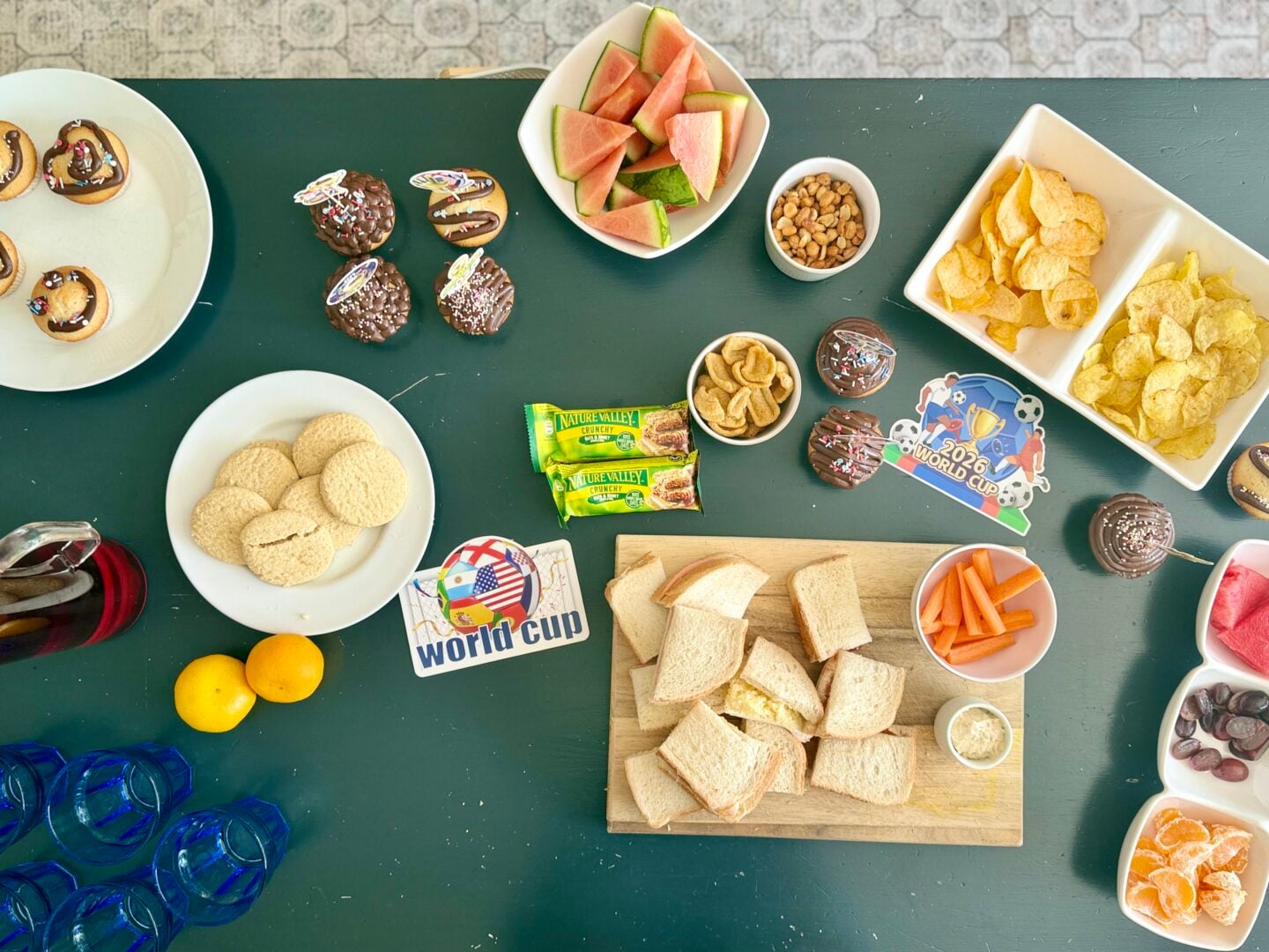 How To Host A Football Watch Party At Home This Summer A table set with various snacks including bread, crisps, biscuits, fruit, sweets, and drinks. “World Cup” stickers and themed items are also present.
