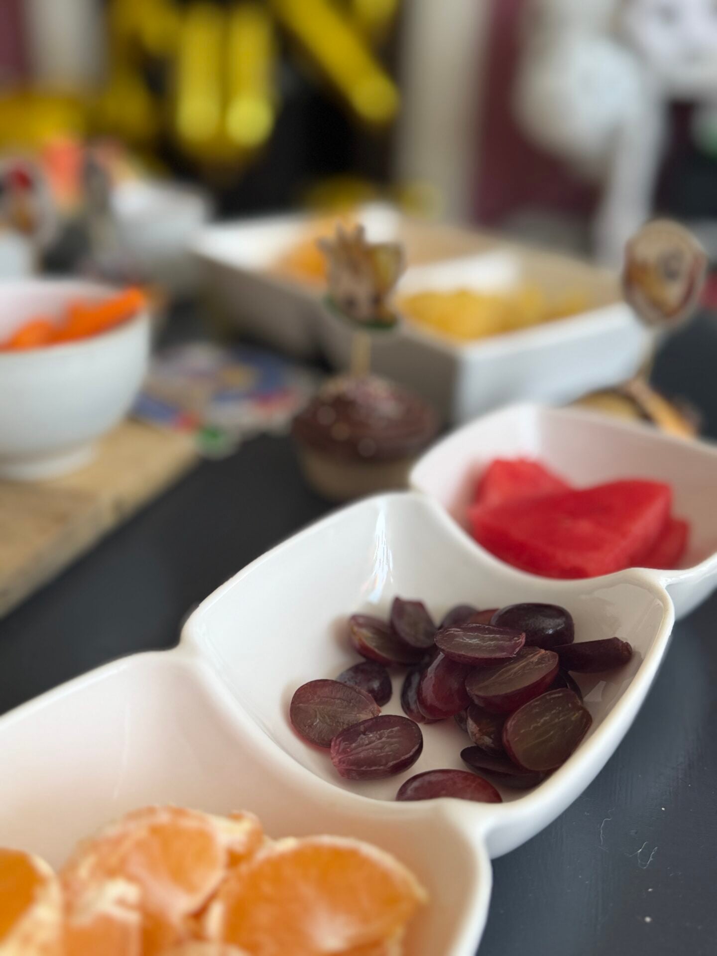 How To Host A Football Watch Party At Home This Summer Close-up of white bowls containing sliced grapes, watermelon, and orange segments on a black table, with more food dishes blurred in the background.