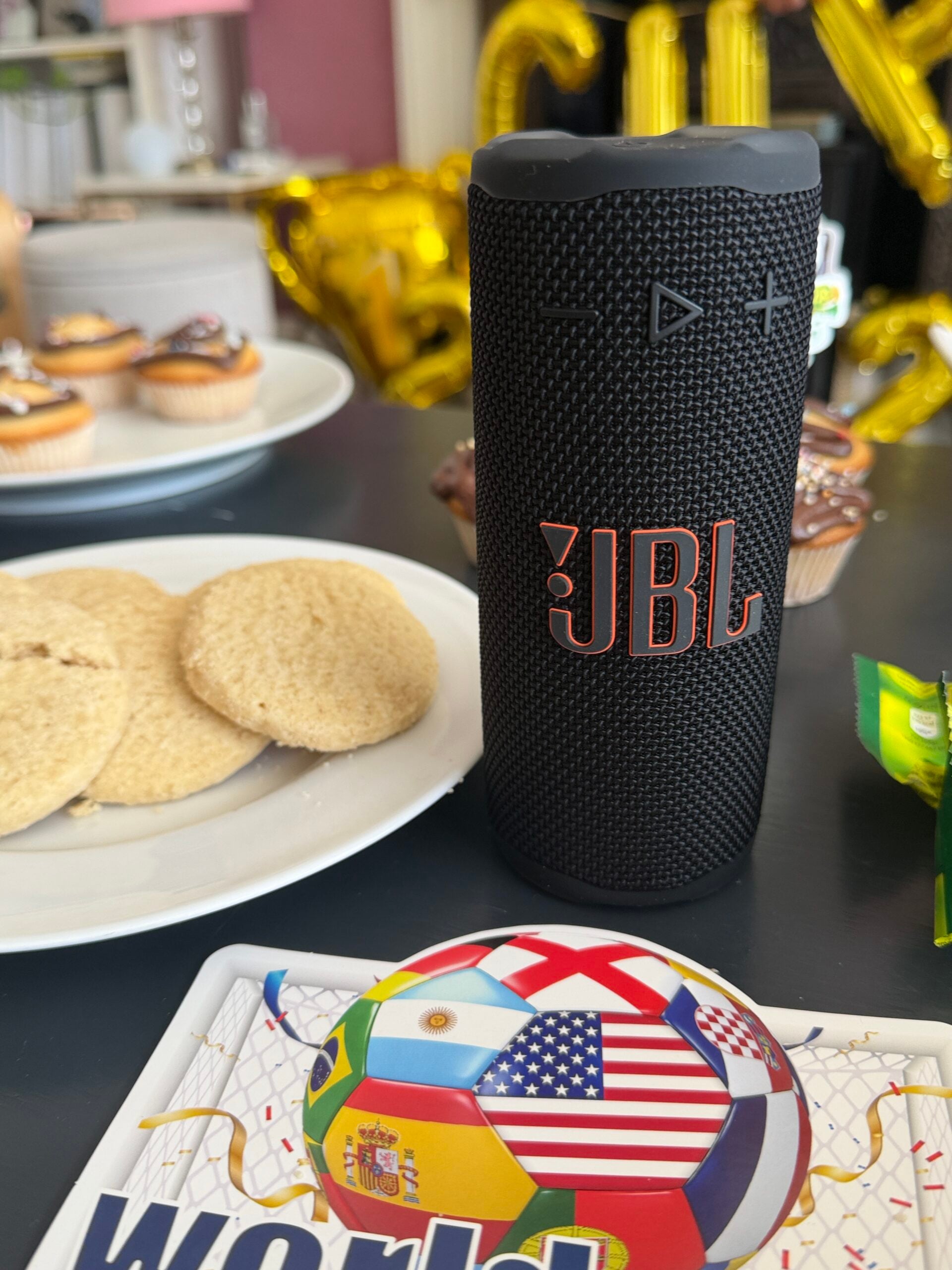 How To Host A Football Watch Party At Home This Summer A black JBL portable speaker sits on a table with biscuits, cupcakes, a packet of sweets, and a World Cup-themed plate in the foreground. Gold balloons are in the background.