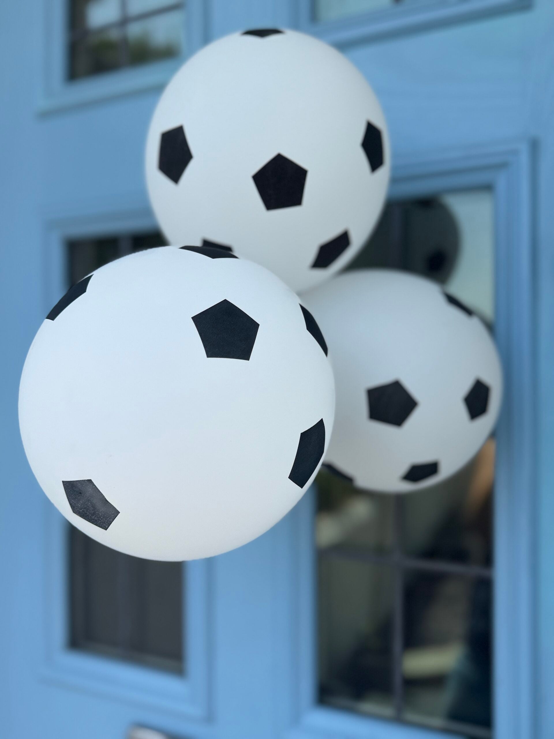 How To Host A Football Watch Party At Home This Summer Three white balloons with black pentagons resembling footballs are floating in front of a blue door with glass panes.