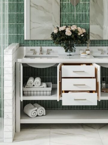 How to Choose a Bathroom Vanity That Actually Survives Family Life Modern bathroom vanity with open cupboards and drawers, neatly arranged towels, baskets, and a vase of flowers on the worktop-perfect for a family bathroom with stylish green tiles and marble surfaces.