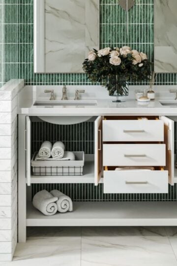 Modern bathroom vanity with open cupboards and drawers, neatly arranged towels, baskets, and a vase of flowers on the worktop—perfect for a family bathroom with stylish green tiles and marble surfaces.