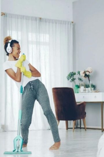 Young woman wearing headphones and rubber gloves pretends to sing into a mop in a bright, tidy room with microcement finishes, with a desk, chair, and plants in the background.