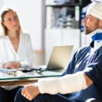 A man with bandages and a neck brace sits at a desk opposite a woman in an office setting, with a laptop and documents visible.