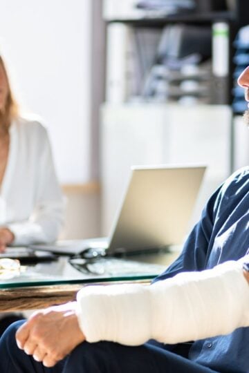 A man with bandages and a neck brace sits at a desk opposite a woman in an office setting, with a laptop and documents visible.