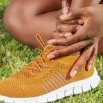 A person sits on grass wearing an orange trainer with a white sole, holding their ankle with both hands.