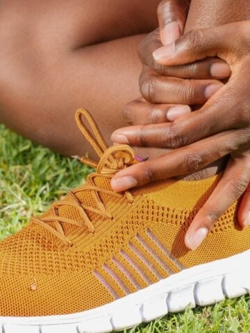 A person sits on grass wearing an orange trainer with a white sole, holding their ankle with both hands.