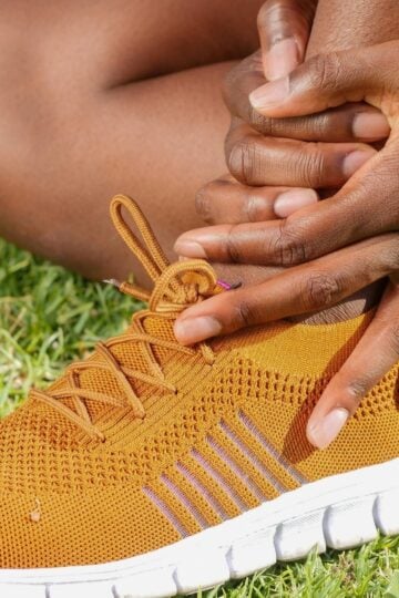 A person sits on grass wearing an orange trainer with a white sole, holding their ankle with both hands.