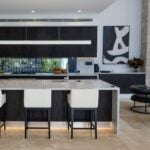 Modern kitchen with a marble island, two white and black bar stools, dark cabinetry, abstract wall art, stylish finishes, and a bowl of fruit on the worktop. Sleek kitchen floor tiles complete the contemporary look.