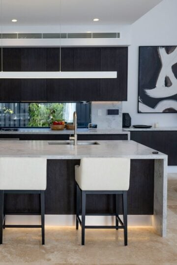 Modern kitchen with a marble island, two white and black bar stools, dark cabinetry, abstract wall art, stylish finishes, and a bowl of fruit on the worktop. Sleek kitchen floor tiles complete the contemporary look.