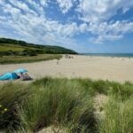Sandy beach with people relaxing under parasols, grassy dunes in the foreground-an ideal spot for a young family. Green hills and blue sky with clouds stretch in the background, capturing the beauty Swansea is known for in any area guide.