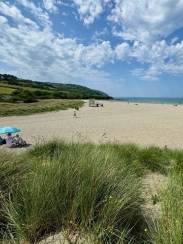 Sandy beach with people relaxing under parasols, grassy dunes in the foreground-an ideal spot for a young family. Green hills and blue sky with clouds stretch in the background, capturing the beauty Swansea is known for in any area guide.