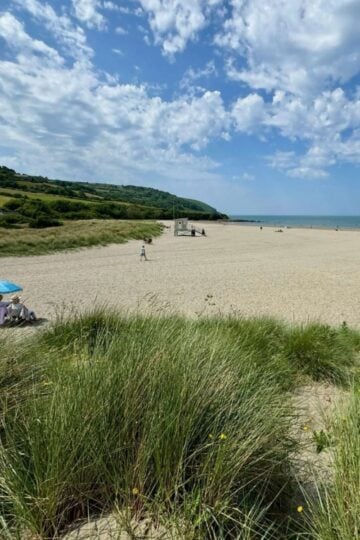 Where to Live in Swansea if You've Got a Young Family: A Welsh Mum's Honest Area Guide Sandy beach with people relaxing under parasols, grassy dunes in the foreground—an ideal spot for a young family. Green hills and blue sky with clouds stretch in the background, capturing the beauty Swansea is known for in any area guide.