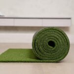 A green artificial grass mat is partially unrolled on a light-coloured floor, with a white cupboard visible in the background-showcasing the ease of installing artificial grass and adding style to any space.