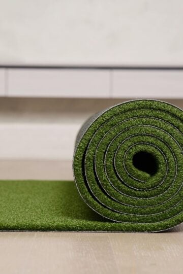 A green artificial grass mat is partially unrolled on a light-coloured floor, with a white cupboard visible in the background—showcasing the ease of installing artificial grass and adding style to any space.