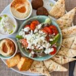 Why Greek Flatbread Adds Versatility To Everyday Cooking A plate with Greek salad topped with feta, pitta bread slices, falafel, houmous, tzatziki, two fried chips, and another dip, all arranged on a white platter.