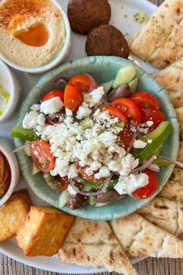 A plate with Greek salad topped with feta, pitta bread slices, falafel, houmous, tzatziki, two fried chips, and another dip, all arranged on a white platter.
