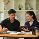 Two people sit at a desk with books, a laptop, and stationery, discussing something on a tablet in a modern office-perhaps exploring hiring tips as they consider whether to hire a personal assistant for their team. Shelves line the background.