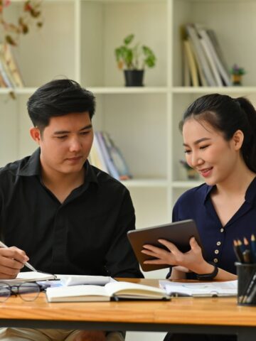 7 Things to Look for When You Hire a Personal Assistant Two people sit at a desk with books, a laptop, and stationery, discussing something on a tablet in a modern office-perhaps exploring hiring tips as they consider whether to hire a personal assistant for their team. Shelves line the background.