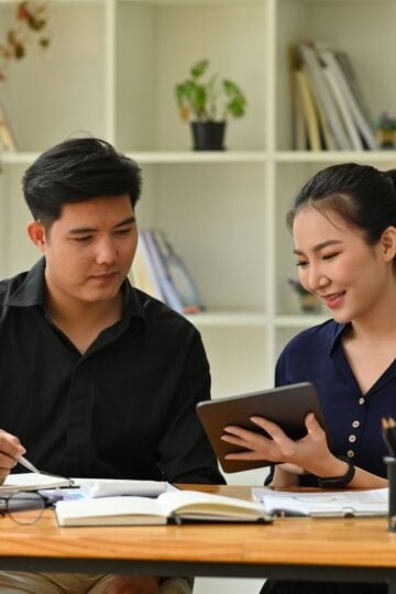 Two people sit at a desk with books, a laptop, and stationery, discussing something on a tablet in a modern office—perhaps exploring hiring tips as they consider whether to hire a personal assistant for their team. Shelves line the background.
