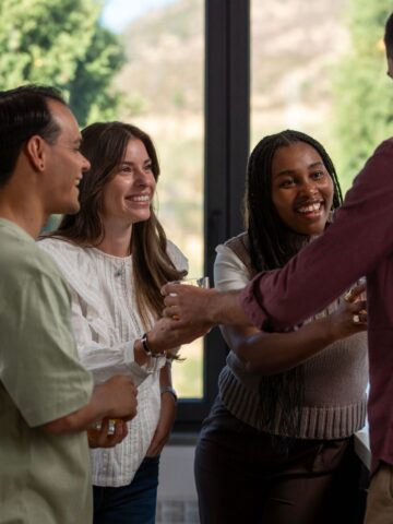 Four people stand indoors near large windows, talking and smiling whilst holding drinks, sharing booking tips for their next social sport experience.