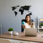 Woman sitting at a desk with a laptop, holding a small globe, with a world map on the wall behind her-perfectly capturing the spirit of global hotel marketing strategy and boosting conversions.