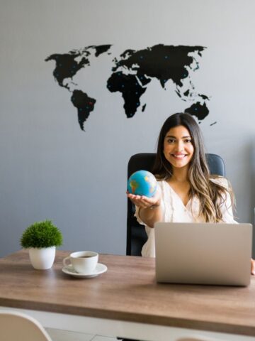 Why Your Hotel Marketing Isn't Driving Enough Conversions Woman sitting at a desk with a laptop, holding a small globe, with a world map on the wall behind her-perfectly capturing the spirit of global hotel marketing strategy and boosting conversions.