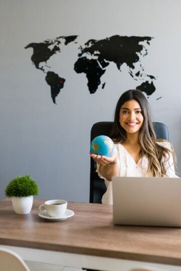 Woman sitting at a desk with a laptop, holding a small globe, with a world map on the wall behind her—perfectly capturing the spirit of global hotel marketing strategy and boosting conversions.