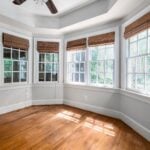 Empty room with wooden floors, three large windows with bamboo blinds, white skirting, and sunlight streaming in-perfect for considering window replacement or evaluating window replacement cost before making updates.