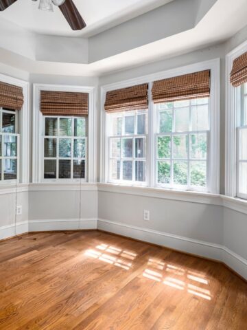 Empty room with wooden floors, three large windows with bamboo blinds, white skirting, and sunlight streaming in-perfect for considering window replacement or evaluating window replacement cost before making updates.