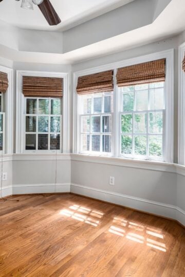 Empty room with wooden floors, three large windows with bamboo blinds, white skirting, and sunlight streaming in—perfect for considering window replacement or evaluating window replacement cost before making updates.