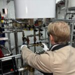 A person wearing gloves adjusts valves on a complex system of pipes and gauges in an industrial setting.