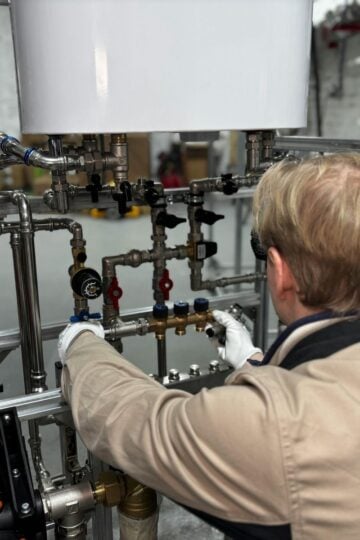 A person wearing gloves adjusts valves on a complex system of pipes and gauges in an industrial setting.