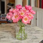 A clear glass vase with pink peonies sits on a round marble table in a room with bookshelves and a colourful crocheted blanket-an inviting scene that's the perfect choice for gifting flowers.