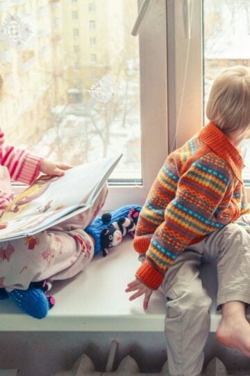 Three children sit on a windowsill; one reads a book whilst the others gaze outside. Cosy in warm, colourful clothes, they show the importance of child safety and creating a secure, childproof home environment.