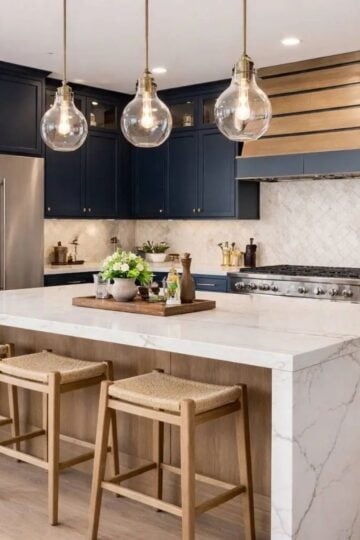 Modern kitchen with navy cupboards, marble island with wooden stools, stainless steel appliances, pendant lights, and neutral splashback.