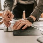 Leather Awl Types: Which One Do You Actually Need for Your Project? Person using a leather awl to cut pieces of black material on a work surface, wearing a watch and a grey jacket, focused on their leather project.