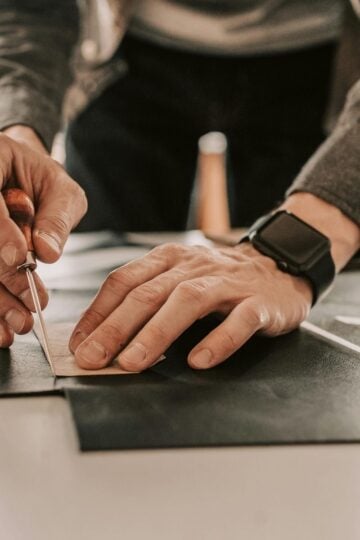 Person using a leather awl to cut pieces of black material on a work surface, wearing a watch and a grey jacket, focused on their leather project.