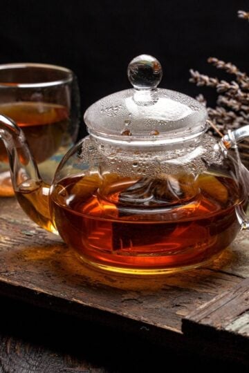 A glass teapot, known for being the healthiest material for making tea, sits on a rustic wooden tray beside a glass cup of tea and dried lavender.