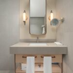 Modern bathroom vanity with a rectangular basin, wall-mounted mirror, two wall lights, a magnifying mirror, and two white towels hanging on a wooden cupboard.