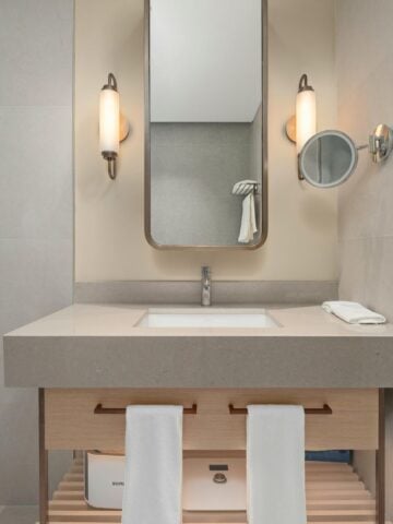 How to Choose the Right Stone Vanity Top Finish? Modern bathroom vanity with a rectangular basin, wall-mounted mirror, two wall lights, a magnifying mirror, and two white towels hanging on a wooden cupboard.